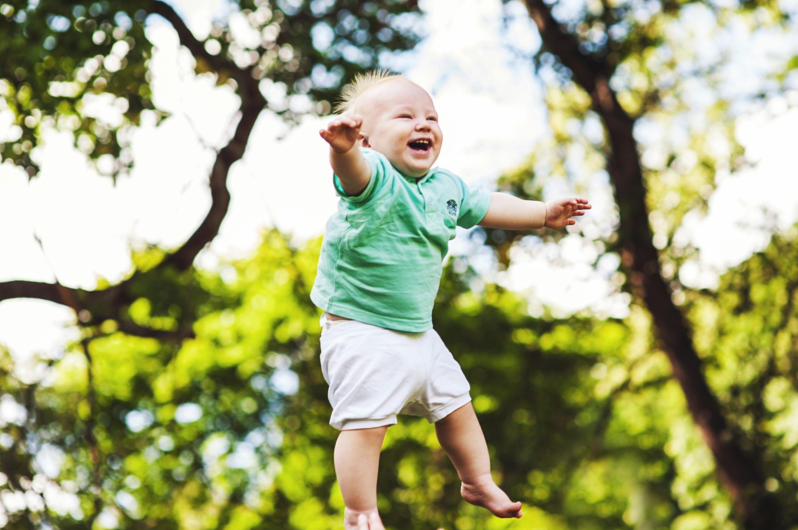Happy baby wearing a green shirt being tossed in the air on a sunny day in the park.