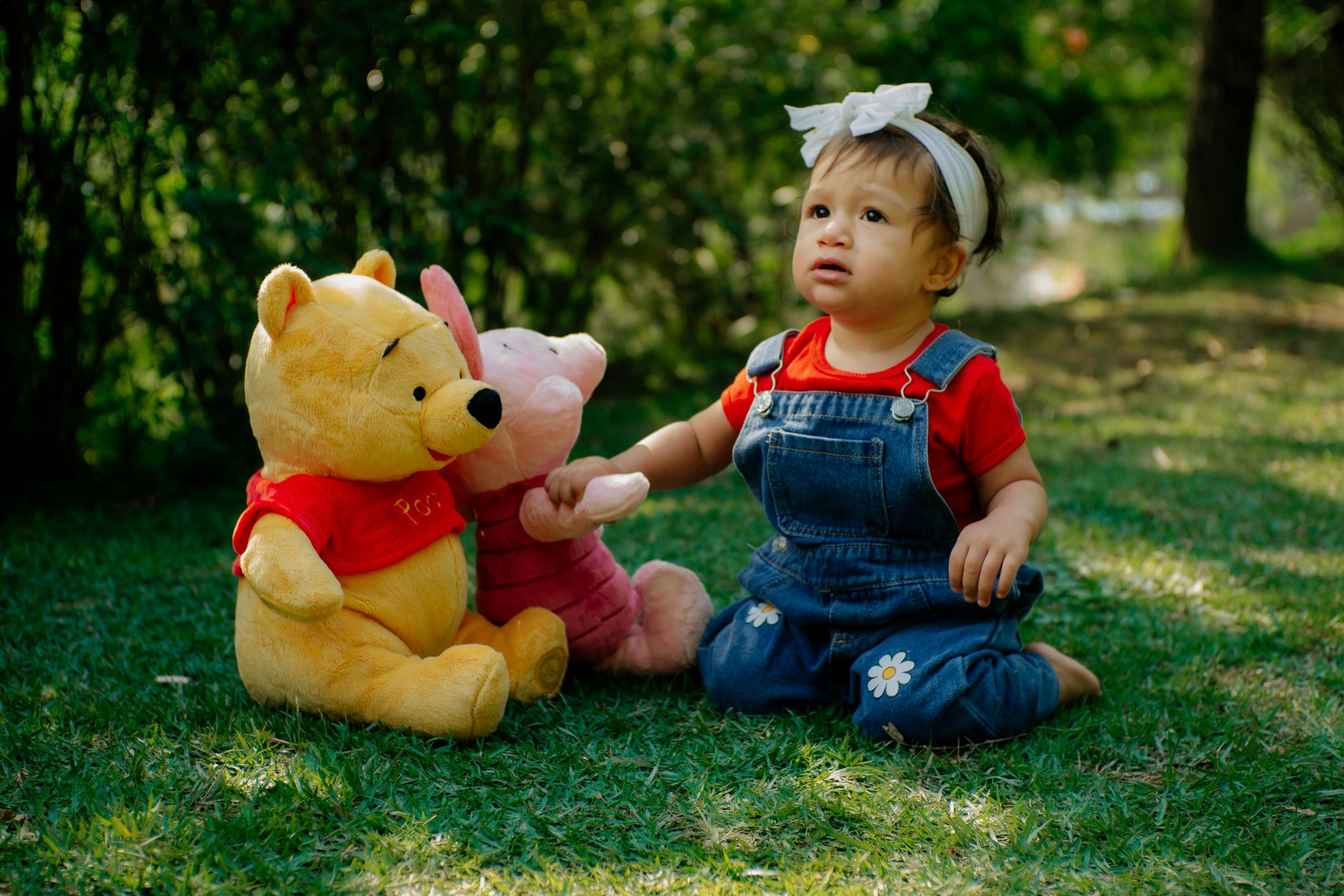 Adorable child in denim and red plays with Pooh Bear plush toys on grass.